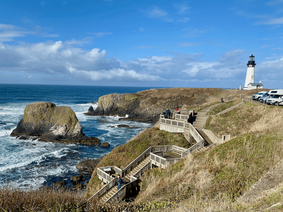 Yaquina Head Lighthouse rises up on a spit of land surrounded by waves crashing against dramatic rock stacks. There are some cars parked right on the edge of the cliff with a staircase leading down to the rocky beach.