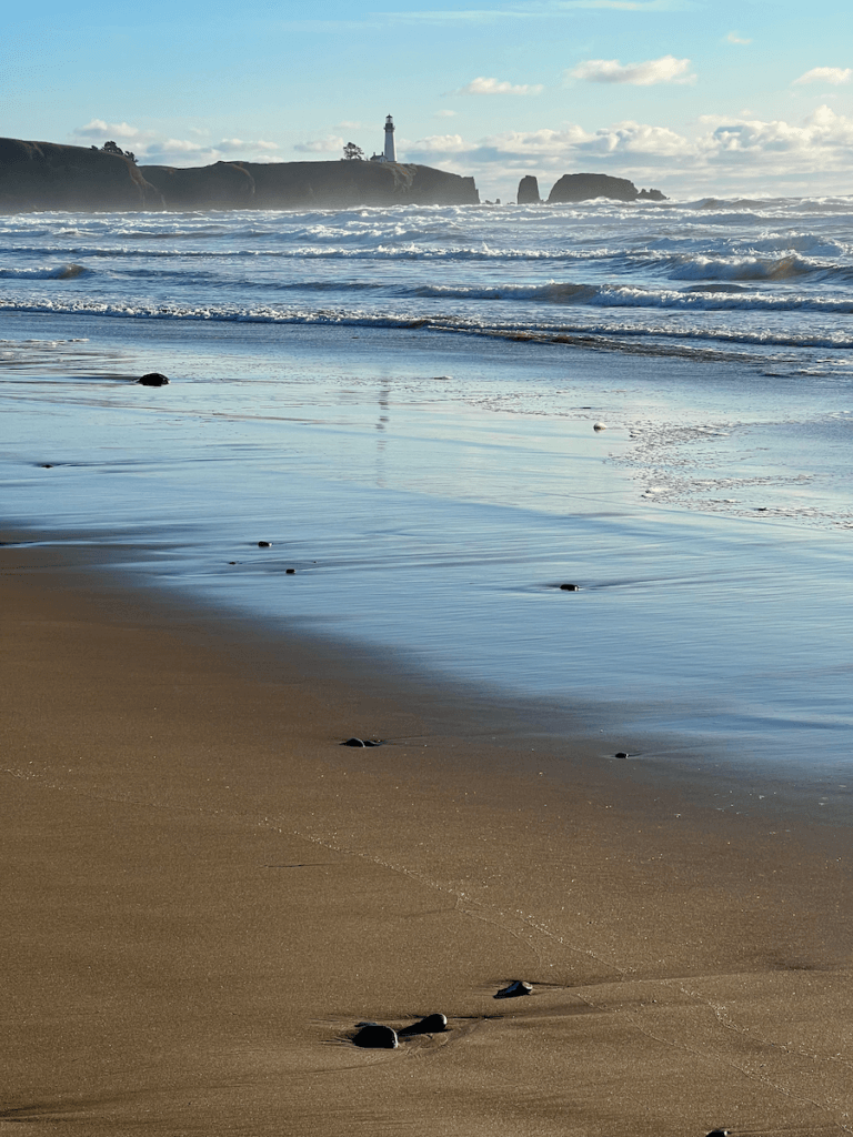 A view of Yaquina Head Lighthouse from a smooth sandy beach. The White and black lighthouse rises up in the distance. This is a wonderful nature thing to do in Newport Oregon.