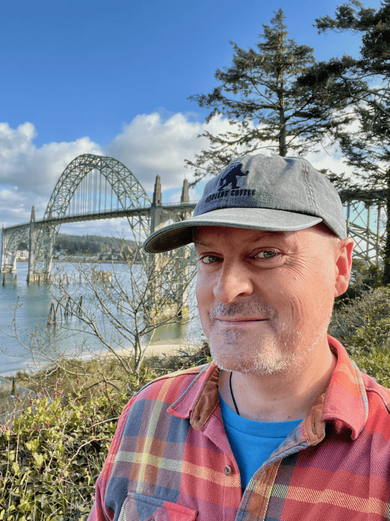 Matthew Kessi poses for a selfie with the Yaquina Bay Bridge in the background in Newport Oreogn. He's wearing an orange plaid shirt and a gray hat and smiling under blue sky with some white clouds.