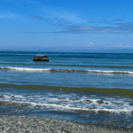 A beach on the Olympic Peninsula in Washington State near a nature immersion retreat center shows the foamy waves washing onto a rocky beach. In the distance there is a large building in the water under a blue sky.