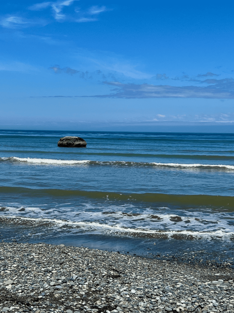 A beach on the Olympic Peninsula in Washington State near a nature immersion retreat center shows the foamy waves washing onto a rocky beach. In the distance there is a large building in the water under a blue sky.
