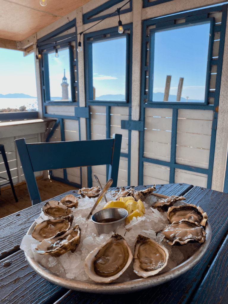 A tray of fresh shucked oysters sit ready to be devoured at Taylor Shellfish in Samish along the Salish Sea. The deck is painted blue and white and you can see the San Juan Islands in the distance under bright blue skies.