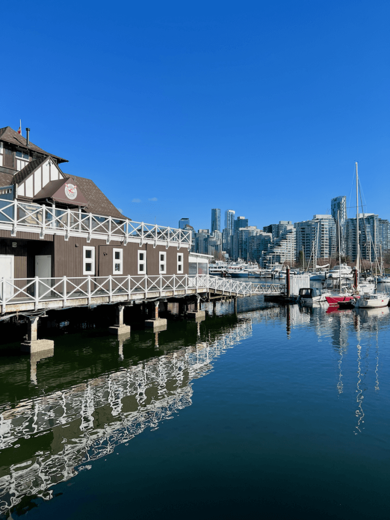 Vancouver British Columbia under bright blue skies. Here, a boathouse drapes over the bay while sailboats park in the moorage. In the background are high rise towers on the styling.