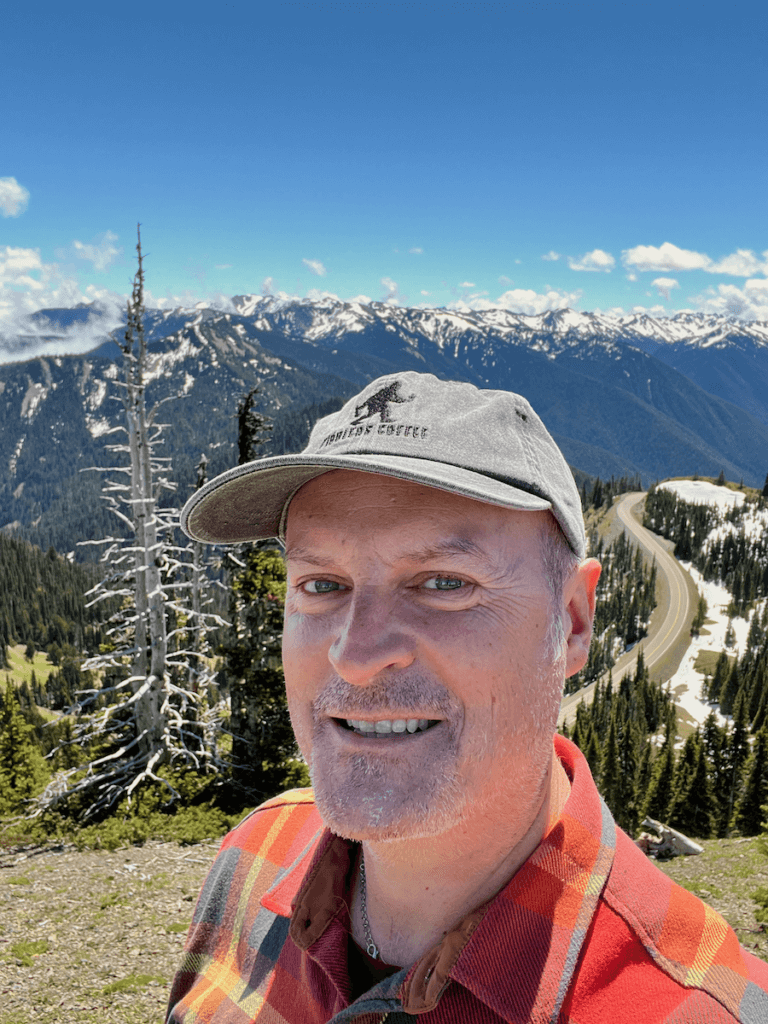 Matthew Kessi poses for a selfie atop Hurricane ridge while on an Olympic Peninsula road trip. The mountains are snow capped in the distance as a windy road weaves along a pass way below in the valley.