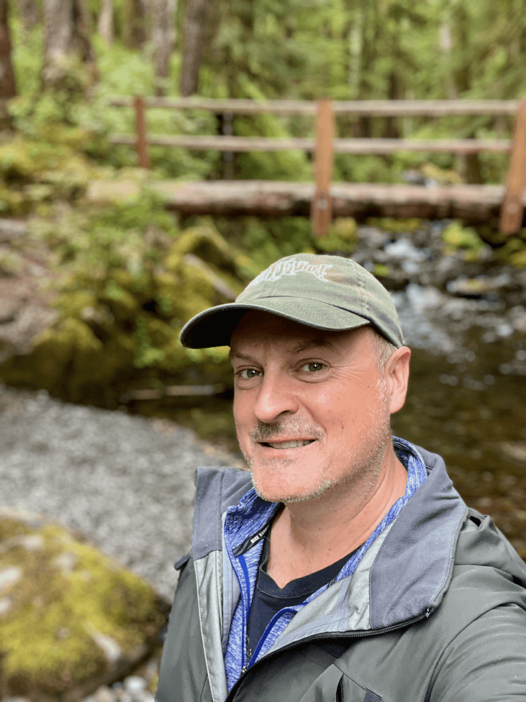 Matthew Kessi stands for a selfie while facilitating a nature immersion retreat on the Olympic Peninsula. He's smiling, wearing a green hat and rain jacket, and there is a bridge crossing a babbling creek in the background.