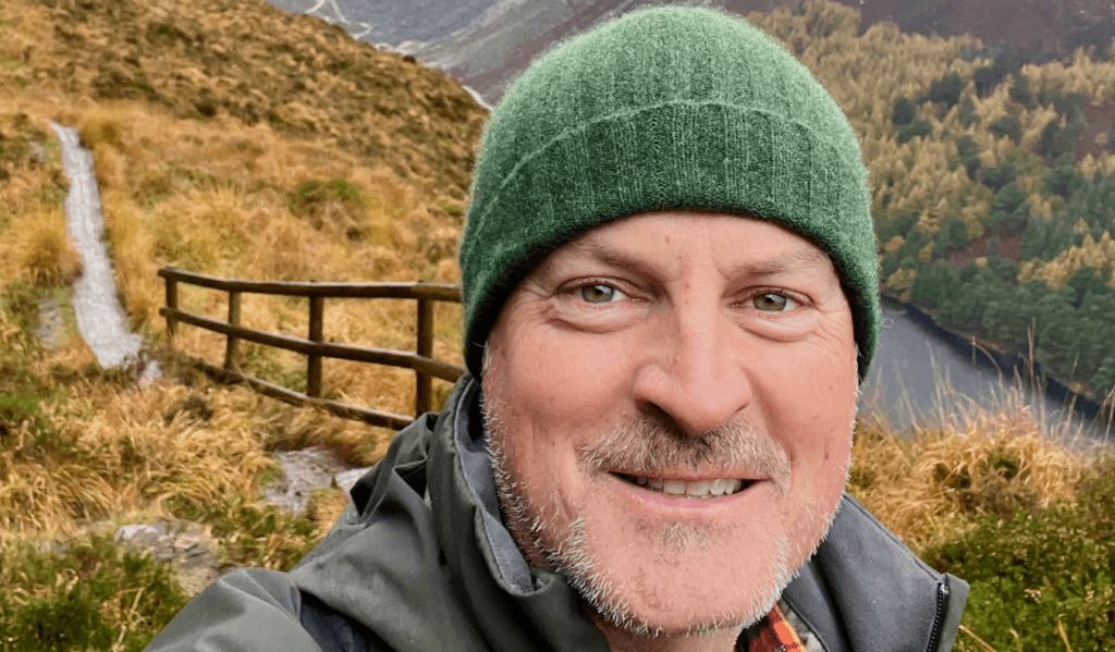 Matthew Kessi selfie on a hike in a fall setting high on a cliff overlooking a placid lake. There is a boardwalk behind him with yellow and red fall colors. Matthew is a practical mystic offering powerful nature meditation coaching programs.