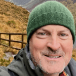 Matthew Kessi selfie on a hike in a fall setting high on a cliff overlooking a placid lake. There is a boardwalk behind him with yellow and red fall colors. Matthew is a practical mystic offering powerful nature meditation coaching programs.