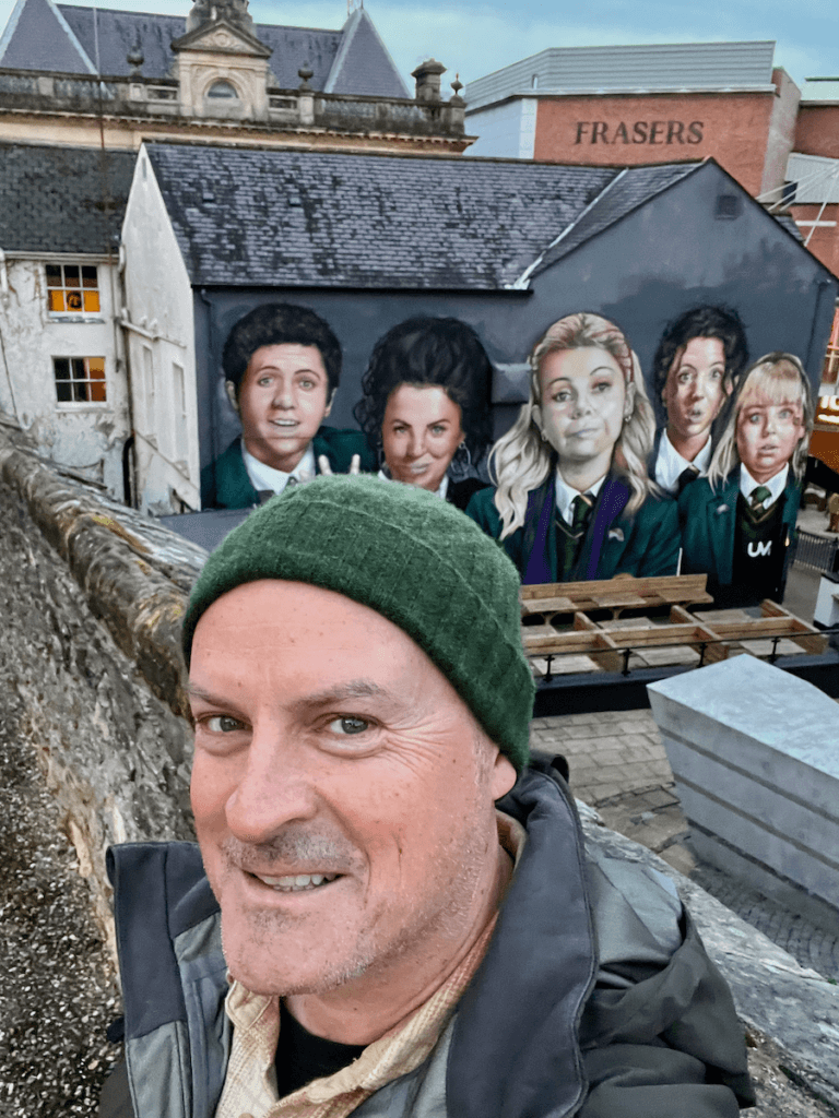 Matthew Kessi stands for a selfie in front of a mural on a wall in the city of Derry in Northern Ireland. The painting depicts the main characters of the hit TV show Derry Girls. He's smiling while wearing a green cap and gray coat.