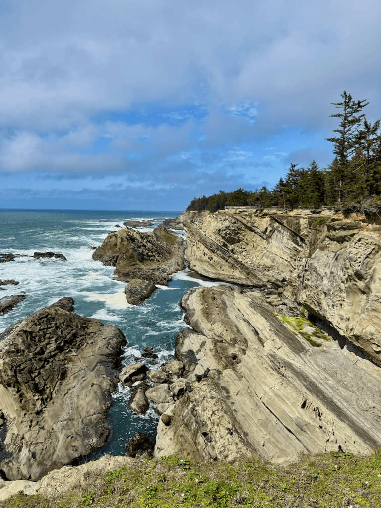 This dramatic shot on the Oregon Coast shows grayish blue water foaming up against sandstone rocks jutting up to the sky in a forty-five degree angle. A good example of how travel planning can set up a wonderful tour of the Pacific Northwest that includes beautiful seascapes like this one with trees along cliffs above the water.