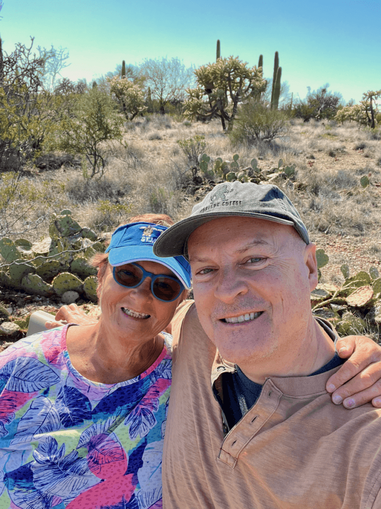 Matthew Kessi and Georgia Kessi take a rest after walking on a trail in Saguaro National Park while experiencing a nature connection experience. She's wearing a brightly colored floral shirt and a blue visor while he's wearing a dust covered t-shirt with a gray hat. There are a variety of cactus in the background under blue skies.