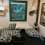 The inside of an eclectic lodge in the Pacific Northwest shows the morning after, with the plaid bed sheets ruffled and two pillows strewn about the bed. The window has a green border and blinds and there is a poster that reads Olympic National Park.