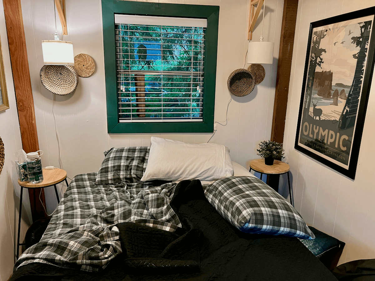 The inside of an eclectic lodge in the Pacific Northwest shows the morning after, with the plaid bed sheets ruffled and two pillows strewn about the bed. The window has a green border and blinds and there is a poster that reads Olympic National Park.
