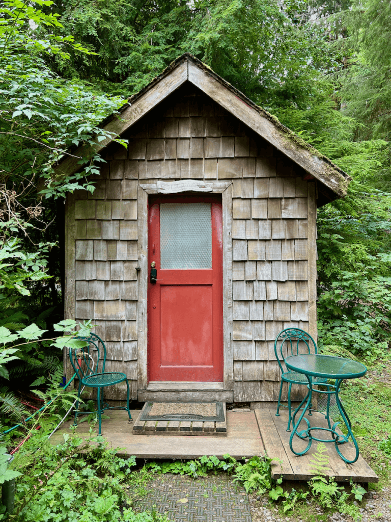 A cute little cabin of a lodge on the Olympic Peninsula is a great place to stay to connect with nature. The door is a dull red with green bistro table and two chairs. The wood cabin is surrounded by cedar and other green shrubbery.