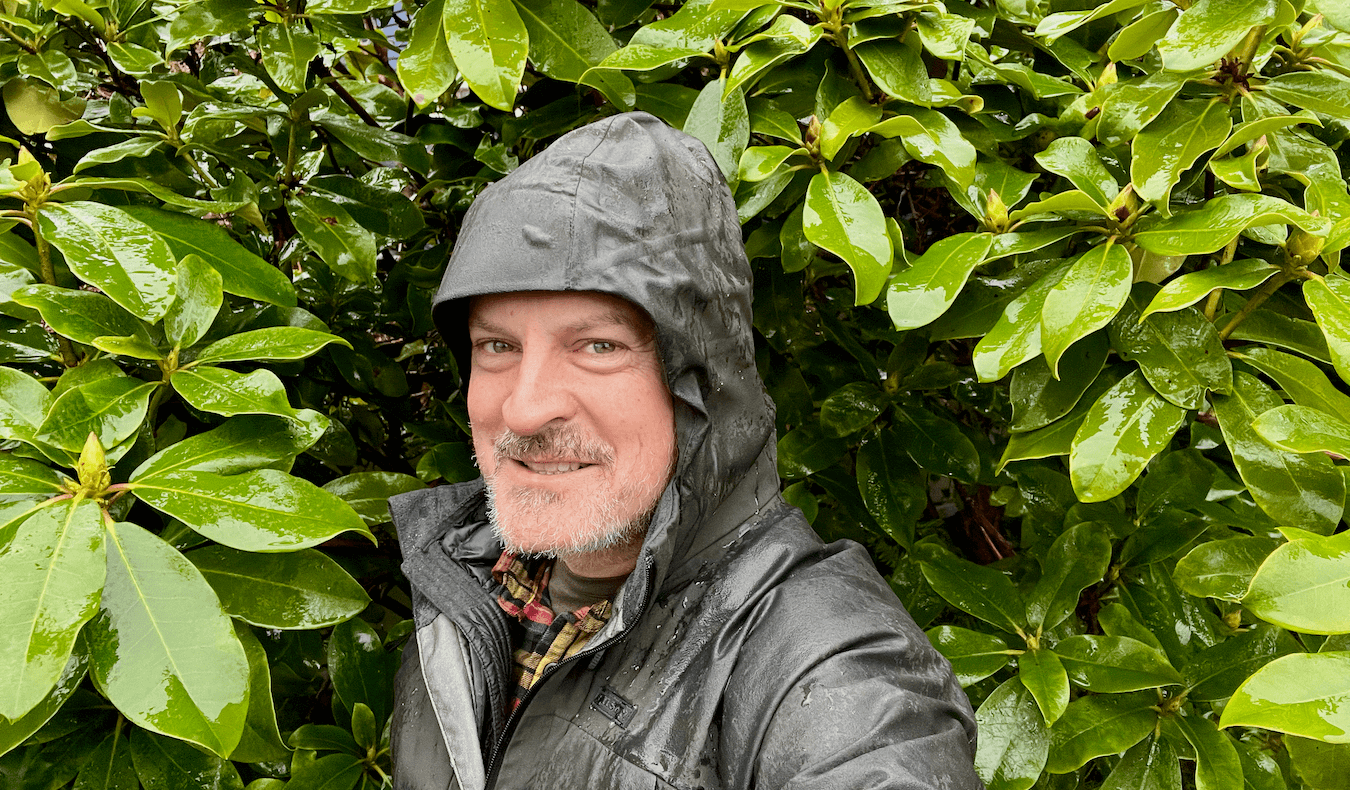 Matthew Kessi poses for a selfie wearing a green raincoat covered in droplets and surrounded by rain-glistening rhododendron leaves. He is a practical mystic helping the world find deeper connections to the Natural World.