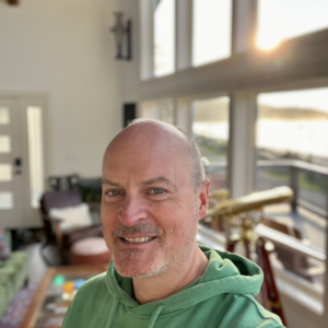 Matthew Kessi smiles from inside a beach house on the Olympic Peninsula in the Pacific Northwest. He is a mystic guide, known for leading people through guided meditation, wellness retreats, and mindfulness coaching. He is smiling big and his head is shaved and he's wearing a green hoodie.