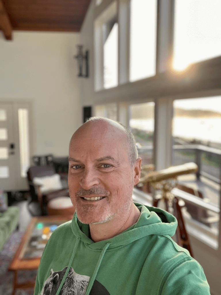 Matthew Kessi smiles from inside a beach house on the Olympic Peninsula in the Pacific Northwest. He is a mystic guide, known for leading people through guided meditation, wellness retreats, and mindfulness coaching. He is smiling big and his head is shaved and he's wearing a green hoodie.