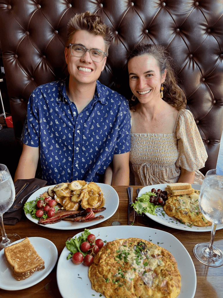 Two eager eaters sit in a booth in downtown Vancouver, Washington waiting to devour pancakes and omelettes and fruit. They are smiling. One is a man with a blue shirt and the other a woman with a cream colored sun dress.