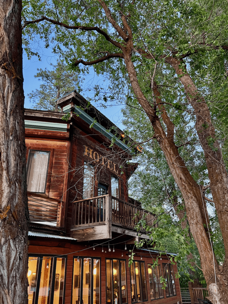 A hotel in Winthrop, Washington is nestled among trees with blue sky above and light glowing in a restaurant on the deck level. 