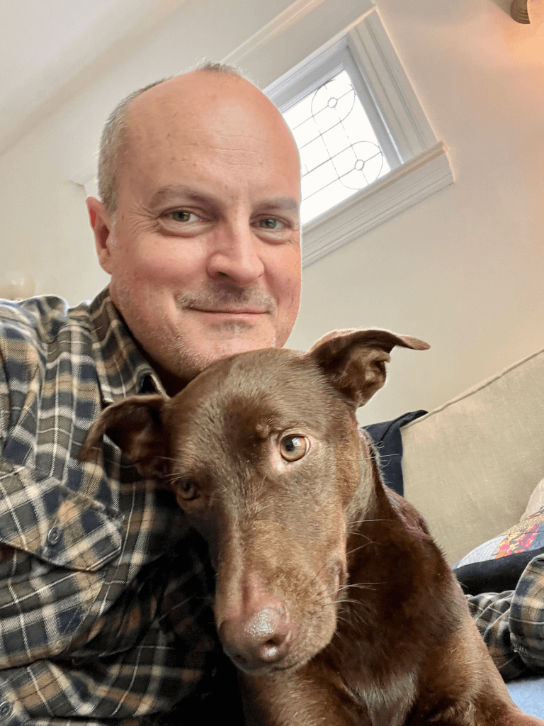 Matthew Kessi smiles as he hugs a brown dog while both of them look into the camera. 