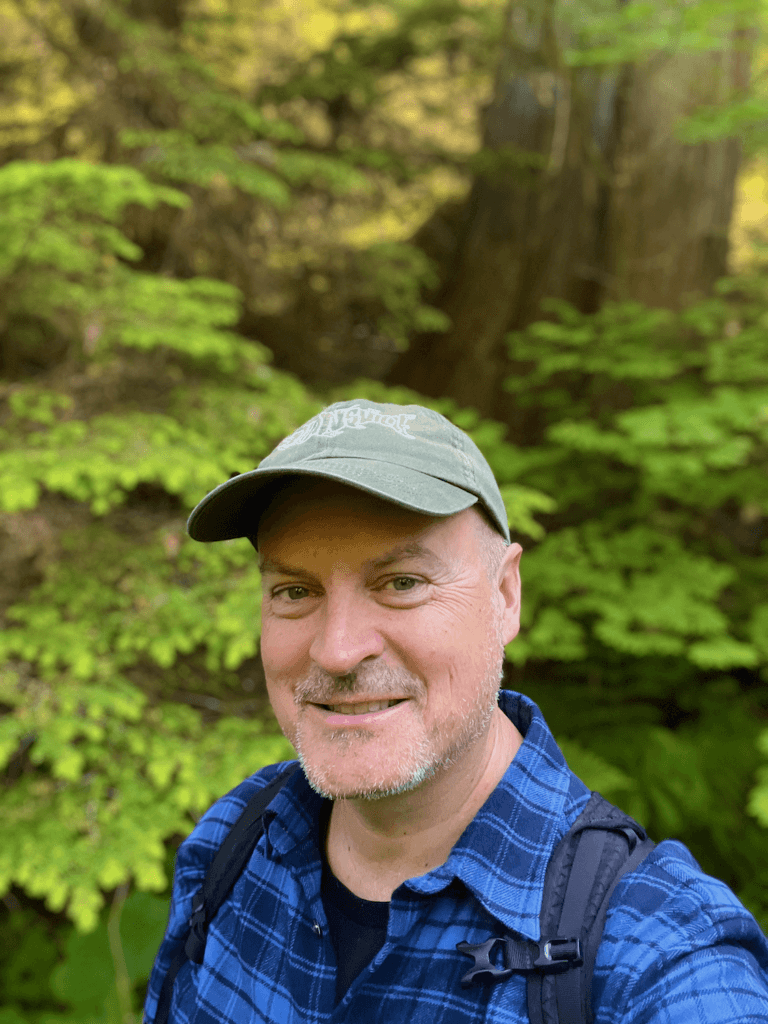 Matthew Kessi poses for a selfie while hiking in North Cascades National park. He's wearing a green cap and a blue plaid shirt with a backpack straps. Bright green foliage and an out of focus tree is in the background.