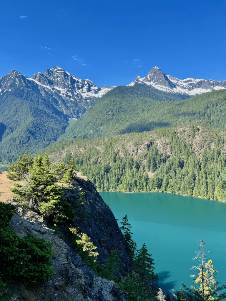 A beautiful view of Diablo Lake on a roadtrip from Seattle to North Cascades National Park. Majestic mountains rise up toward a blue sky, covered in summer snow while hills roll down to a blueish green lake. This shot is taken from a viewpoint on a high vista.