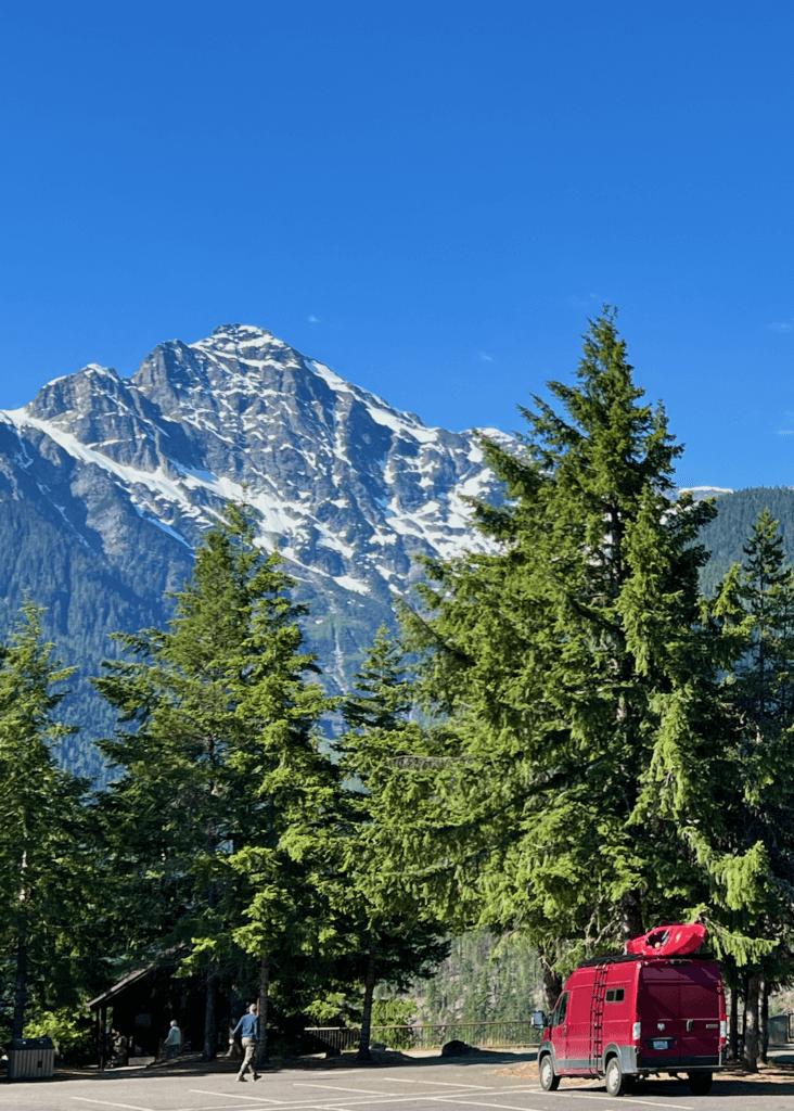 A viewpoint along an epic road trip from Seattle to North Cascades National Park, in Washington State. Jagged mountains still covered in snow rise up into a bright blue sky while green fir trees tower over a red ram travel van with a red kayak on top. A man walks to the railing of the viewpoint.