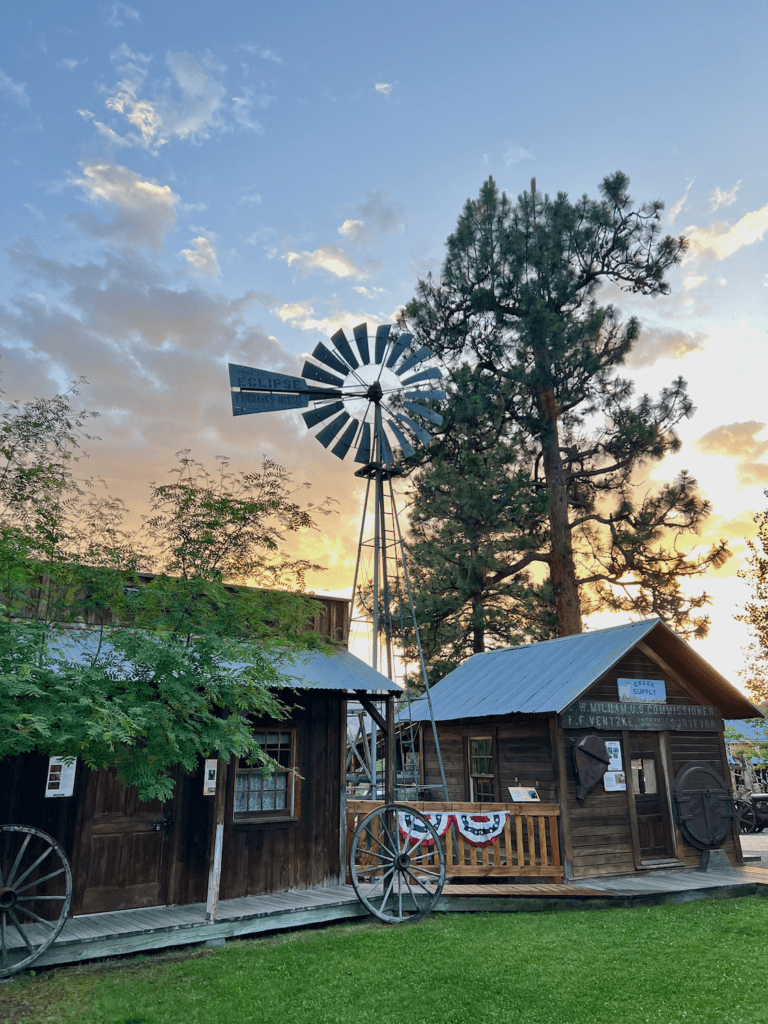 A pioneer scene in Winthrop, Washington makes a fun stop on a road trip from Seattle. There is a windmill and pioneer motif buildings with a lawn of bright green grass in the forefront. The sunset sky is various shades of yellow and blue with puffy clouds.