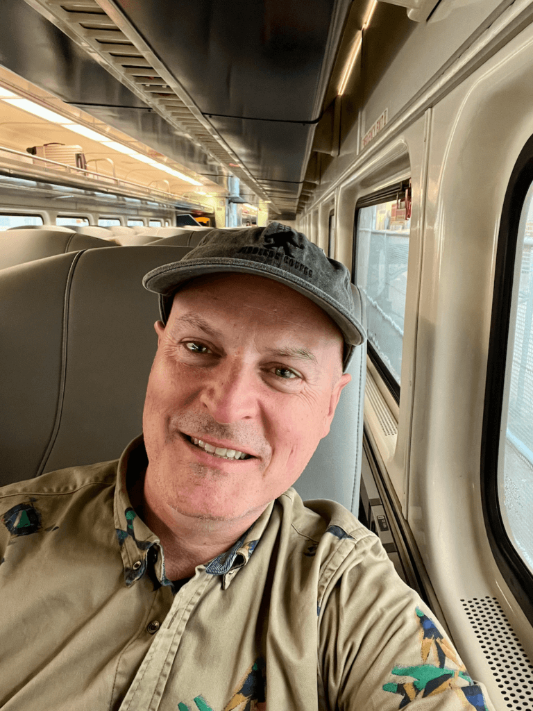 Matthew Kessi smiles while on an Amtrak train going from Seattle to Portland. He's wearing a cap and a multi-colored brown shirt. 