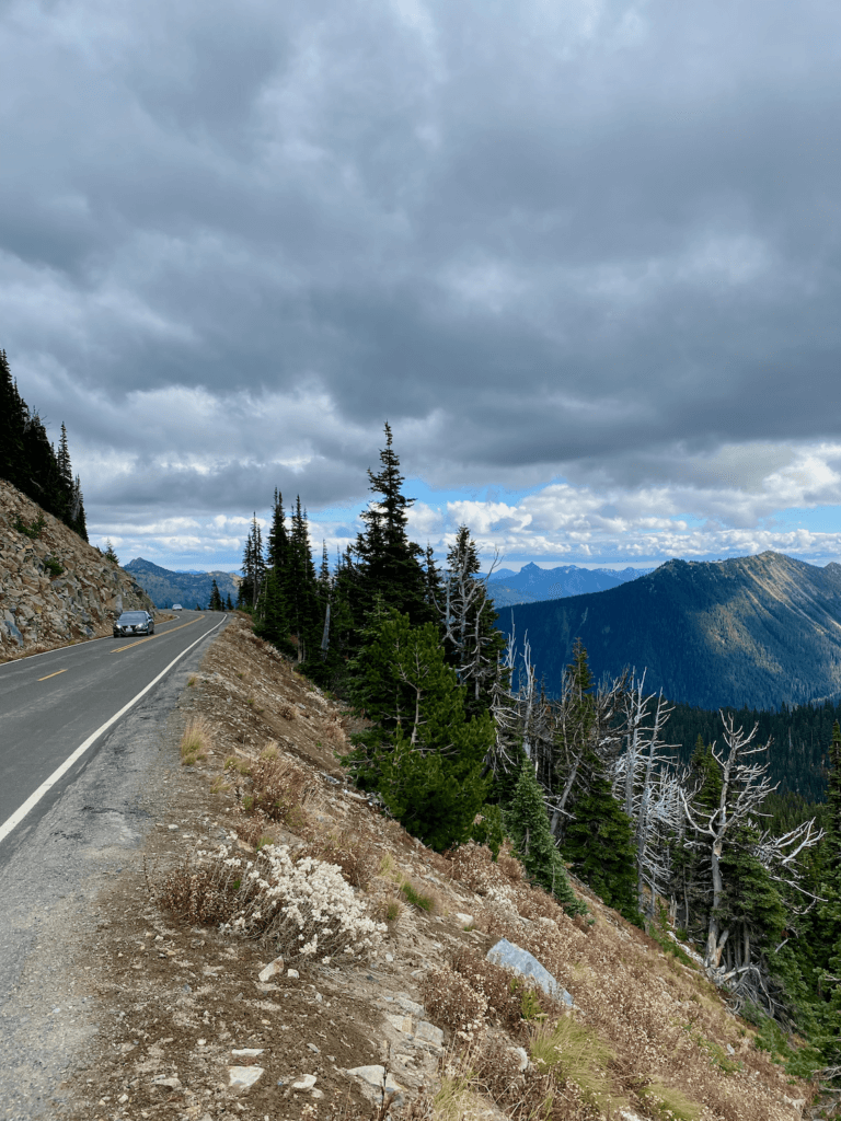 A view from a road on the way from Seattle to Mount Rainier shows a car driving in the distance. There is a curve in the road moving away toward the horizon of gentle mountains under gray cloud cover that appears like it might rain.