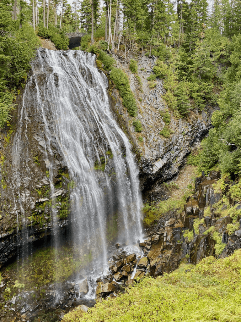 The beautiful cascading waters of Narada Falls on the road from Seattle to Mt Rainier makes for a peaceful stop along the road trip. In the far distance you can see a stone bridge crossing over the river. The foreground has bright green grass and a rocky step down to the misty waterfall.