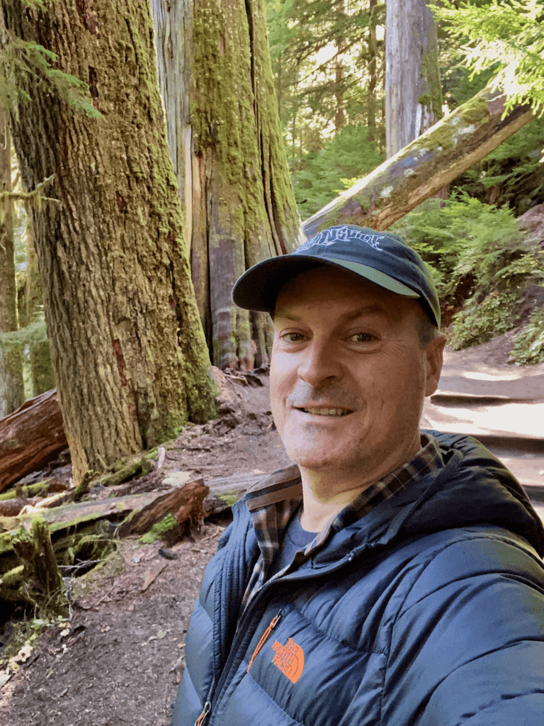 Matthew Kessi poses for a selfie near Ohanapecosh Visitor Center in Mt Rainier National Park. He's in front of some very large and very old trees, smiling. He's wearing a green hat and a gray puffy coat.