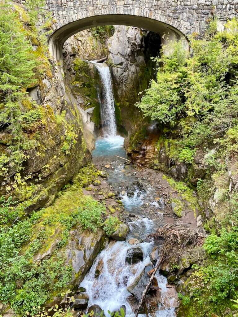 A beautiful waterfall flows under a bridge on the drive from Seattle to Mt. Rainier. The water is prisinte and flowing over glossy rocks and green trees.