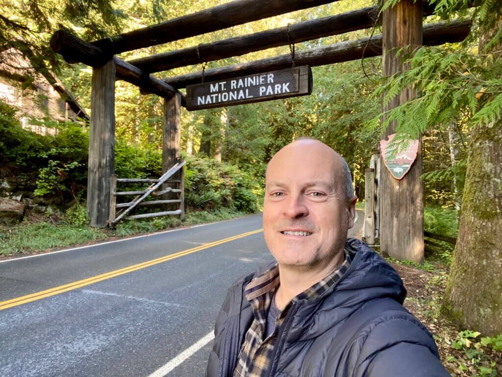 Matthew Kessi stands for a selfie in front of the entrance to Mt. Rainier National Park on a trip from Seattle. He's smiling and wearing a gray puffy coat and plaid brown shirt while the yellow stripe of the pavement glides by in the background. A wooden sign swings above him saying "Mt. Rainier National Park."