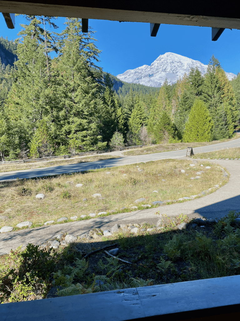 A view of Mt Rainier from the porch of National Park Inn. There are a variety of evergreen trees and rocks in the foreground while the mountain proudly rises up to greet the sky.