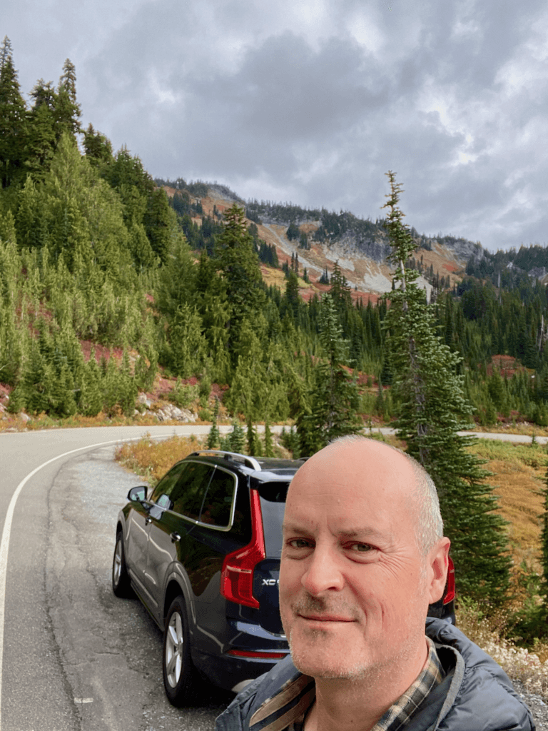 Matthew Kessi poses for a selfie on the side of the road on a tour of Mt. Rainier National Park. He's smiling in font of a blue SUV parked on the shoulder while the hills in the background show changing fall colors of reds and oranges and yellow amidst small evergreen alpine trees.