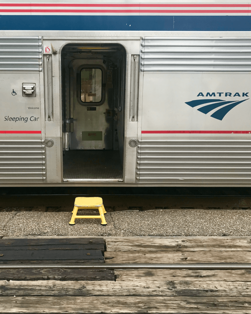 The sleeping car of an Amtrak train going from Seattle to Portland. The outside is shiny and there is a yellow step stool to reach the first floor of the train. 