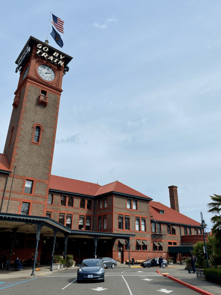 The red brick of Union Station in Portland, Oregon rises up to a clock tower with flags of Oregon and the United States. The sky is blue and cars can be seen driving to pick up passengers. 