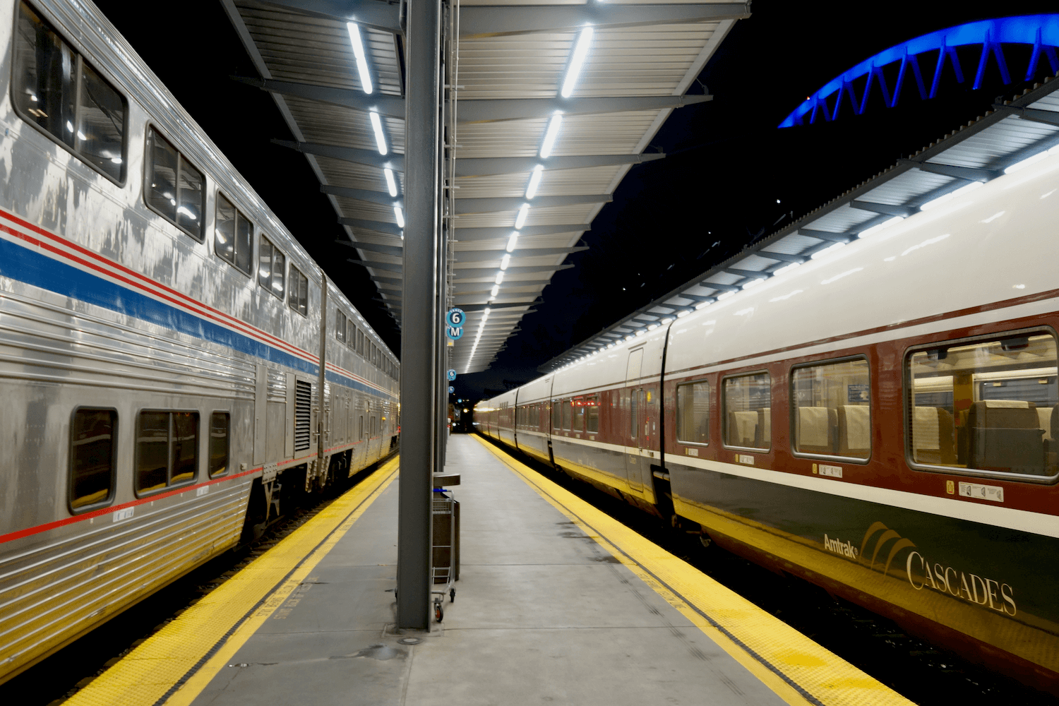 Amtrak operates two kinds of trainsets on the Seattle to Portland train, as shown in this photo. On the left is a double-decker long haul train, with shiny silver siding with blue and red stripes and on the other side is a one-level glossy train with brown and green striping. This photo is taken at the Seattle train station at night.