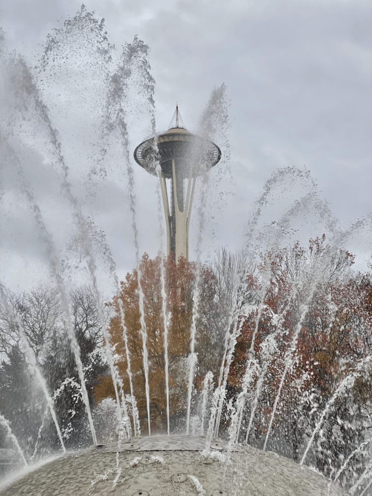 A view of the Space Needle from the International Fountain at Seattle Center. The trees reflect changing fall leaves while jets of water shoot up into the gray sky.