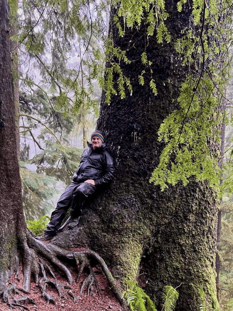 Matthew Kessi poses for a selfie leaning against a tree while forest bathing. He's soaking wet with a thick raincoat and a multi-colored beanie. The sitka spruce is giant while hemlock branches lightly drape in front of the photo. The winding roots of another smaller hemlock tree wind into the earth.