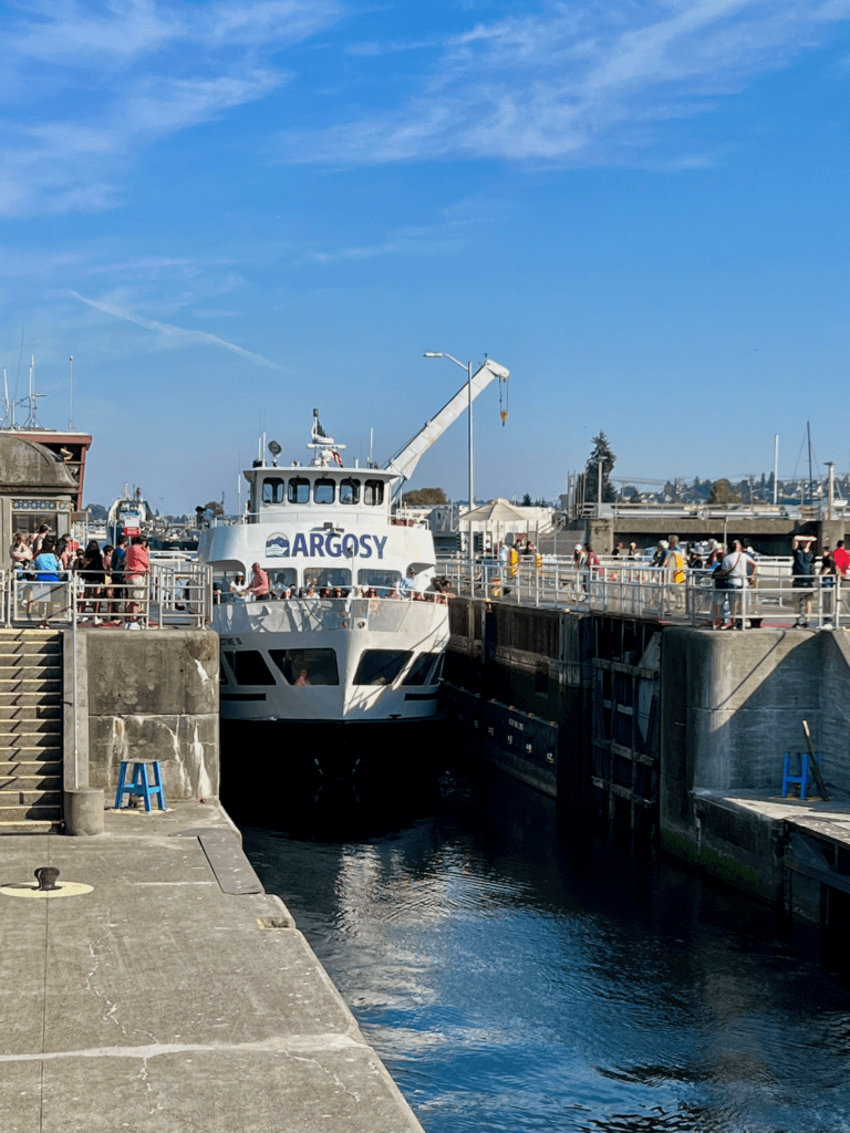 An Argosy Cruise ship navigates through the Ballard Locks in Seattle. The boat has tourists on the deck while people visiting the locks look on from the concert decks. The sky above is blue with slight white clouds.