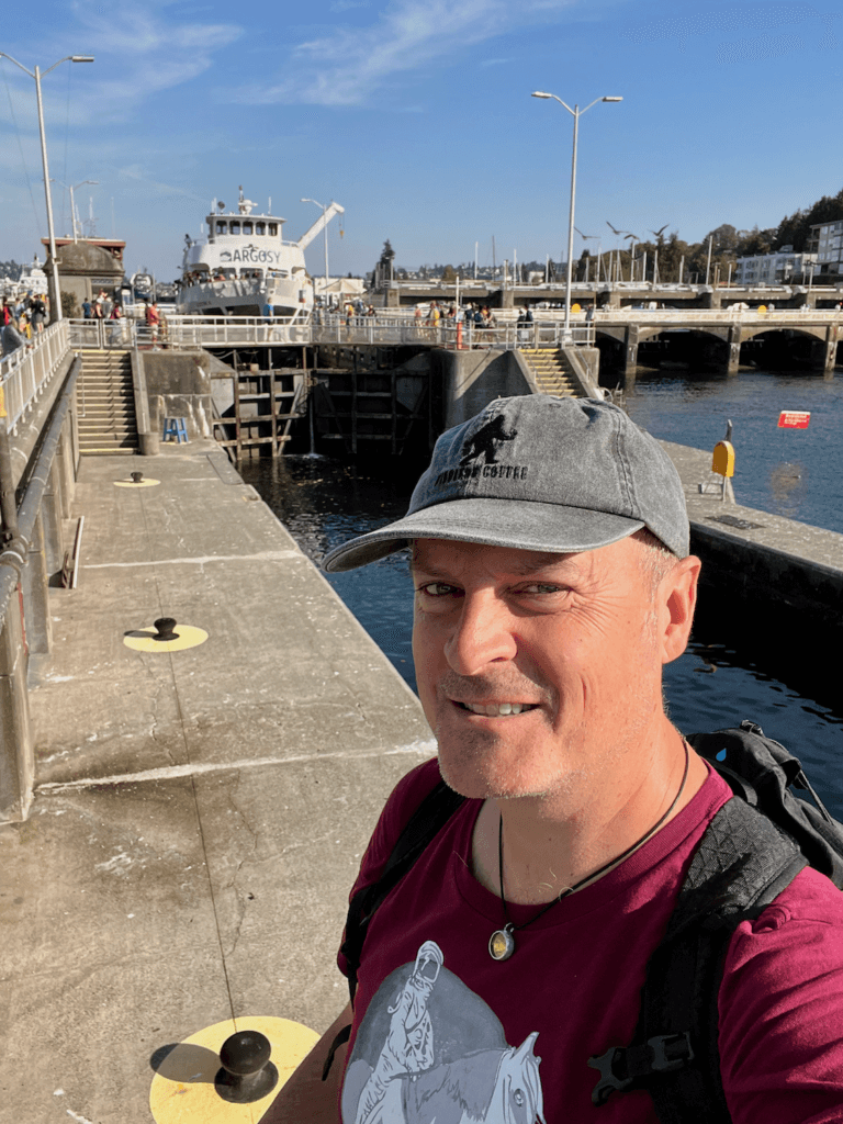 Matthew Kessi poses for a selfie in front of an Argosy Cruises boat going through the Ballard Locks in Seattle. He's wearing a gray hat and purple t-shirt and smiling.