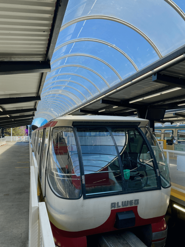 The Seattle Monorail waits at the station near the Space Needle. The futuristic but also retro design of the train, which says Allege on the front. Above the train is a rounded skylight revealing blue sky above.