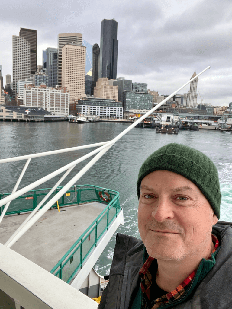 Matthew Kessi poses on the deck of a Washington State Ferry leaving downtown Seattle. The skyline of buildings, including the black Columbia Tower and the iconic Smith Tower, are in the distant background.