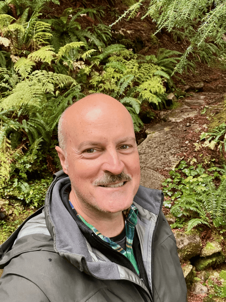 Matthew Kessi smiles while on a nature connection experience in a secret corner of Mt. Rainier National Park. He's wearing layers of gray and green clothing and the forest path behind him is full of different shades of green, and abundant varieties of ferns and moss.