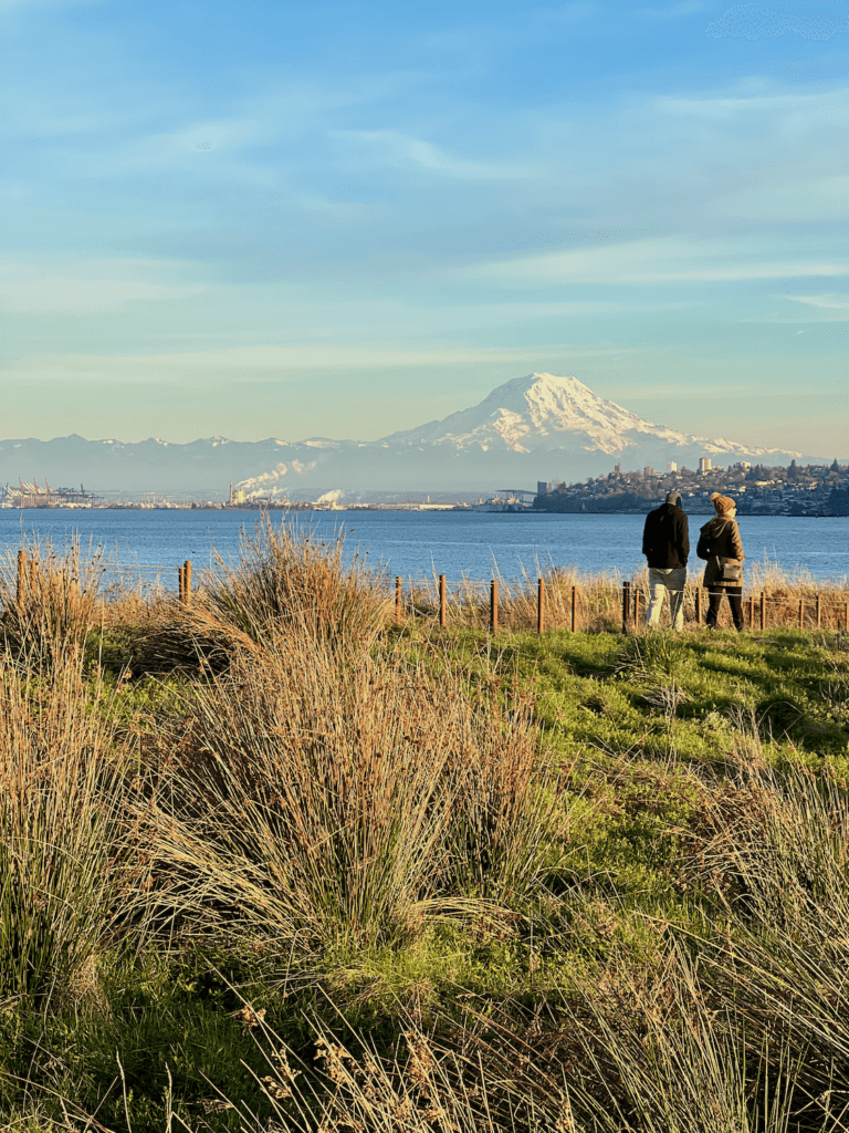 Mt. Rainier rises up majestically above Tacoma, which is a great stop on the drive between Seattle and Portland. Two people walk on a waterfront pathway with the city in the background under blue sky.