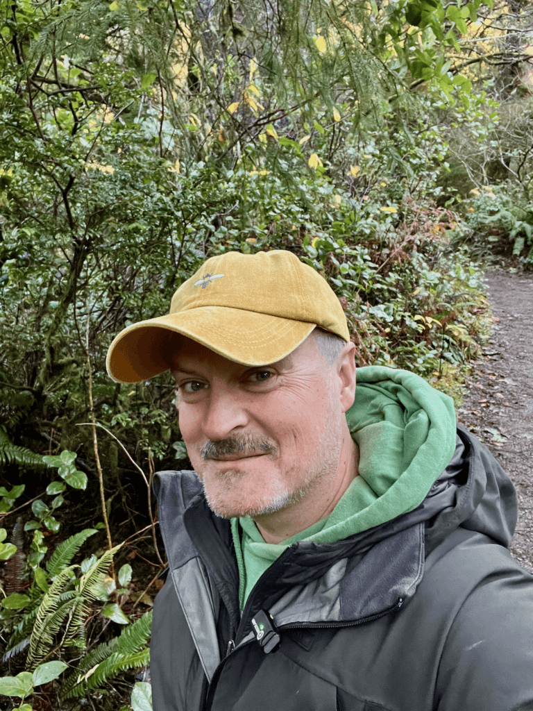 Matthew Kessi walks through Oswald West State Park on the way to a nature connection experience. He's wearing a yellow cap and green hoodie with a gray raincoat. The foliage on the bushes and trees gives a nod to the fall season.