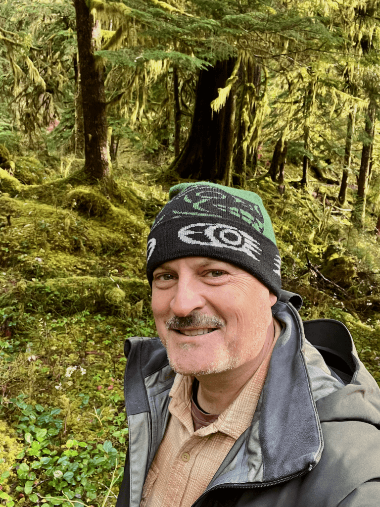 Matthew Kessi smiles while on the Quinault Loop Trail. He's wearing a green and black beanie and a cream colored shirt under a green raincoat. The electric greens of the moss in the background is stunning in contrast.