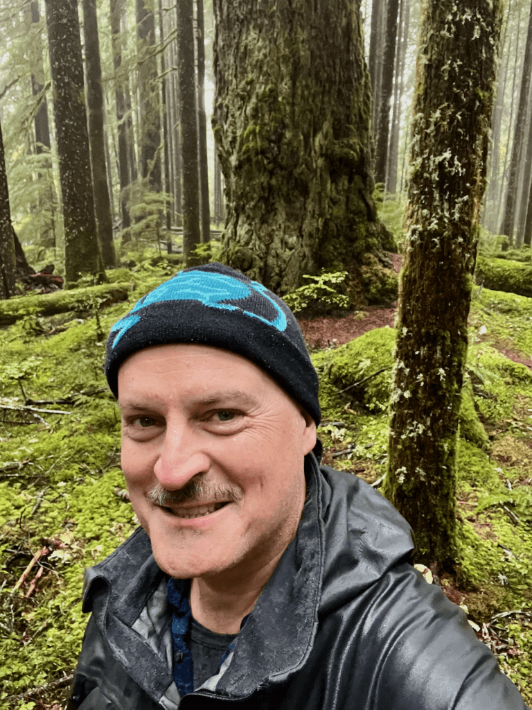 Matthew Kessi smiles for a selfie during a forest bathing adventure deep in a rainforest. The forest floor is covered in thick green moss and a proud ancient Douglas fir rises up behind him.