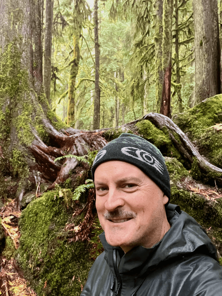 Matthew Kessi smiles for a selfie during a shinrin yoku adventure deep in a rainforest. The forest floor is covered in thick green moss and a proud ancient Douglas fir rises up behind him.